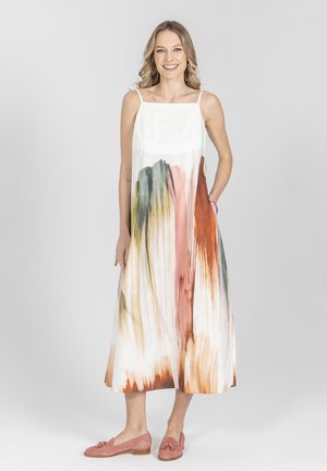 Kobieta uśmiechnięta, stojąca, ubrana w białą, bezrękawnikową sukienkę midi z abstrakcyjnymi wzorami pociągnięć pędzla w kolorach brązowym, zielonym i różowym oraz różowe mokasyny.