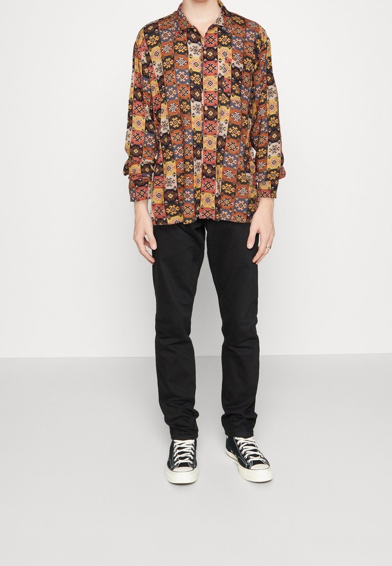 Personne portant une chemise à manches longues à motif géométrique multicolore, un pantalon noir et des baskets noir et blanc, debout devant un fond blanc uni.