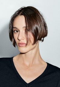 Una donna con un taglio di capelli bob liscio e indossante un top nero a V guarda leggermente di lato, con uno sfondo neutro.