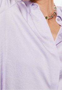 Chemise boutonnée lavande avec une texture lisse et légère, dotée d'un col cranté et d'une légère brillance. Collier de perles avec des accents verts.