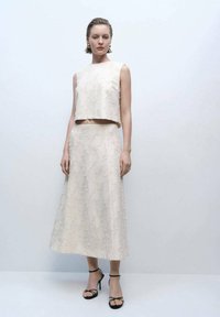 Ensemble crème en deux pièces comprenant un top court et une longue jupe, fabriqué dans un tissu texturé avec des motifs subtils. Talons hauts noirs à brides.