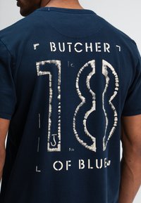Navyblauwe katoenen t-shirt met grote, verouderde witte opdruk "BUTCHER 18" en "OF BLUE" op de achterkant; ronde hals en korte mouwen.