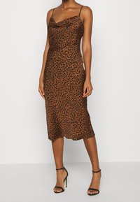 Vestido midi de estampado de leopardo en tonos marrones, diseño con escote asimétrico, tirantes delgados y una textura suave similar a la seda. Combinado con tacones negros con tiras.