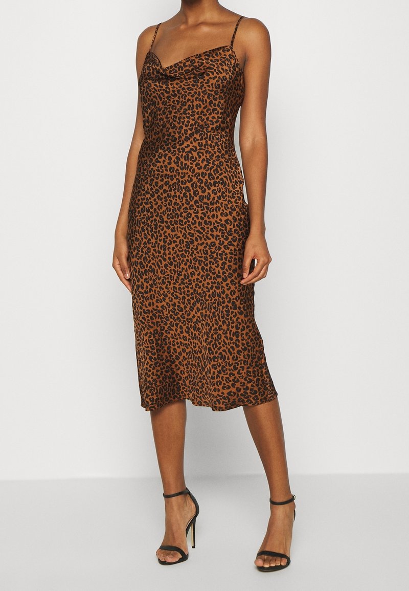 Vestido midi de estampado de leopardo en tonos marrones, diseño con escote asimétrico, tirantes delgados y una textura suave similar a la seda. Combinado con tacones negros con tiras.