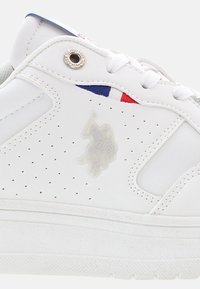 Zapatilla blanca de cuero con paneles laterales perforados, que presenta un logo embosado translúcido y un acento de tela roja y azul en la lengüeta.