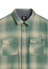 Flannel shirt in green and beige check pattern, featuring a collar, two chest pockets, and metallic buttons. Tag displayed inside collar.