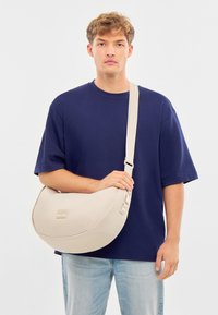 Béžová ramenná taška so semi-kruhovým dizajnom, mäkkou textúrou, nastaviteľným popruhom a malým kovovým logom na prednej strane.