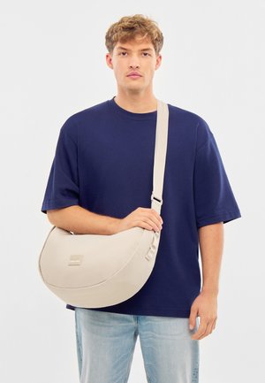 Béžová ramenná taška so semi-kruhovým dizajnom, mäkkou textúrou, nastaviteľným popruhom a malým kovovým logom na prednej strane.