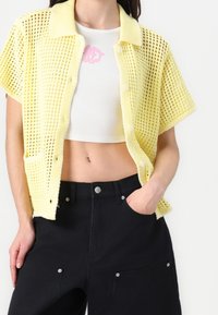 Osoba v žlutém krátkém kardiganu s otevřeným pletením přes bílý crop top s růžovým detailem, doplněná o černé kalhoty s vysokým pasem.
