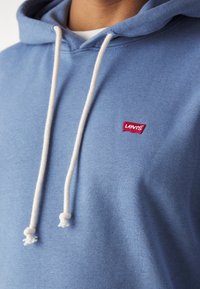 Gros plan d'une personne portant un sweat à capuche Levi's bleu clair avec des cordons blancs et un petit logo rouge Levi's sur la poitrine.