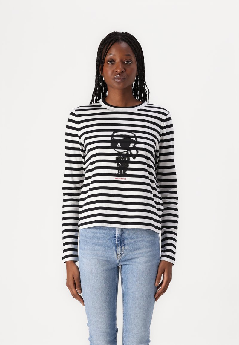 Svart-og-hvitt-stripete langermet skjorte med et sentralt svart grafikk og røde bokstaver. Brukt med lyseblå figursydde jeans.