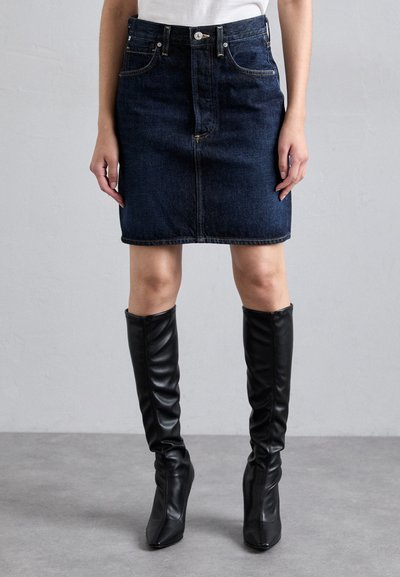 Denimnederdel i mørkeblå med en tætsiddende pasform, to forlommer og synlig søm. Sammen med knæhøje sorte støvler med glat tekstur.