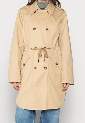 Trench-coat beige croisé avec cordon à la taille et poches à rabat boutonnées, porté sur un top gris et un jean bleu.