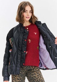 Czarna kurtka puchowa o pikowanej fakturze, z czerwonymi guzikami i podszewką w kwiatowy wzór. Nałożona na czerwoną bluzę z haftowanymi detalami.