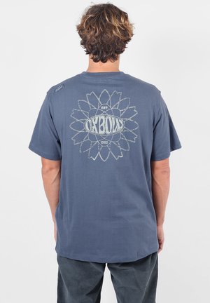 Personne aux cheveux bouclés vue de dos portant un t-shirt bleu avec un motif floral et le texte "OXBOW" dans le dos.