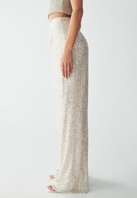 Pantaloni in paillettes di un luccicante colore oro chiaro, con una silhouette dritta e una vita aderente. Accenti di paillettes riflettenti su tutta la superficie.