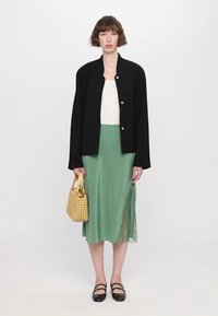 Zwarte knoopjasje over een witte top, gecombineerd met een vloeiende groene rok met een doorzichtige inzet. Accessoires omvatten een gele geweven tas en zwarte schoenen.