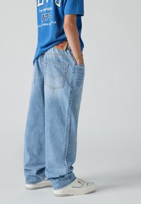 Vaqueros de denim azul claro con un corte relajado, que cuentan con bolsillos traseros y costuras distintivas. Combinados con una camiseta gráfica azul y zapatillas blancas.