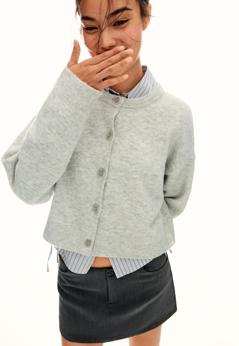 Ung kvinna i grå knappad tröja över randig skjorta och svart kjol, som täcker sin mun med handen medan hon ler.