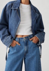 Tummansininen farkkutakki vetoketjulla, ylipitkä malli ja suuri kaulus, yhdistettynä korkeavyötäröisiin vaaleansininisiin farkkuihin.