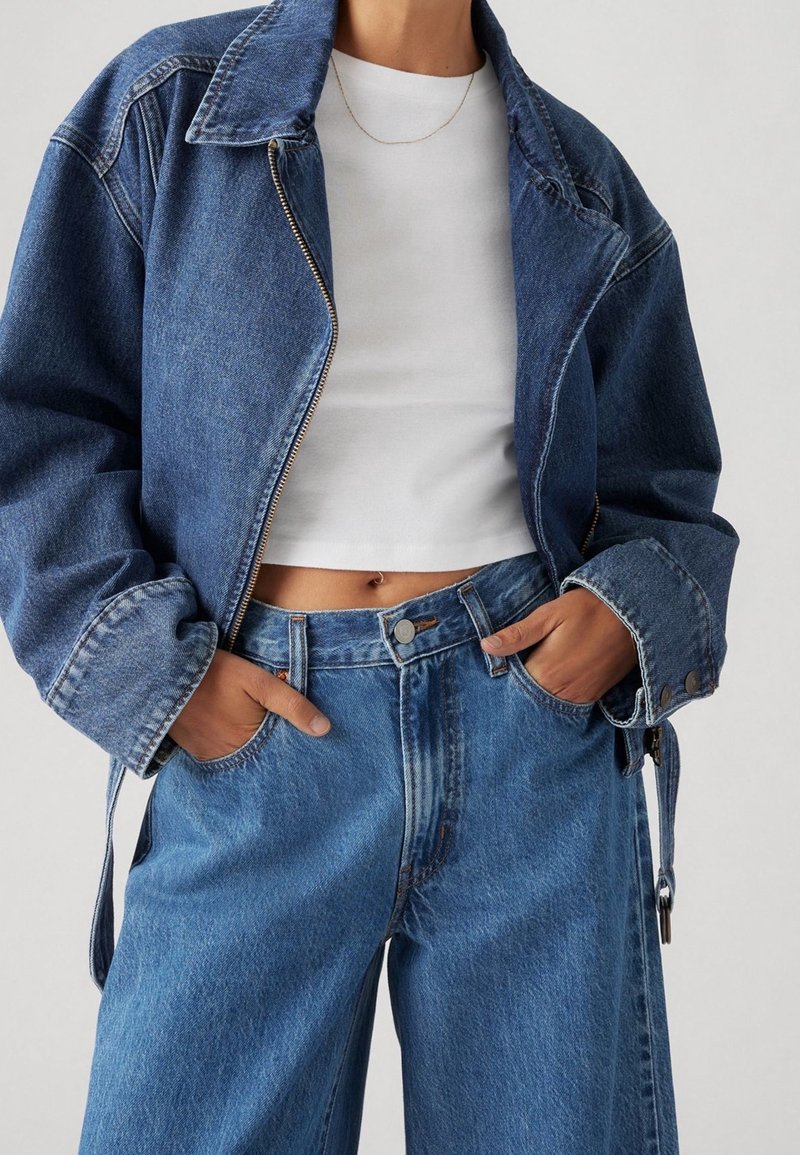 Tummansininen farkkutakki vetoketjulla, ylipitkä malli ja suuri kaulus, yhdistettynä korkeavyötäröisiin vaaleansininisiin farkkuihin.