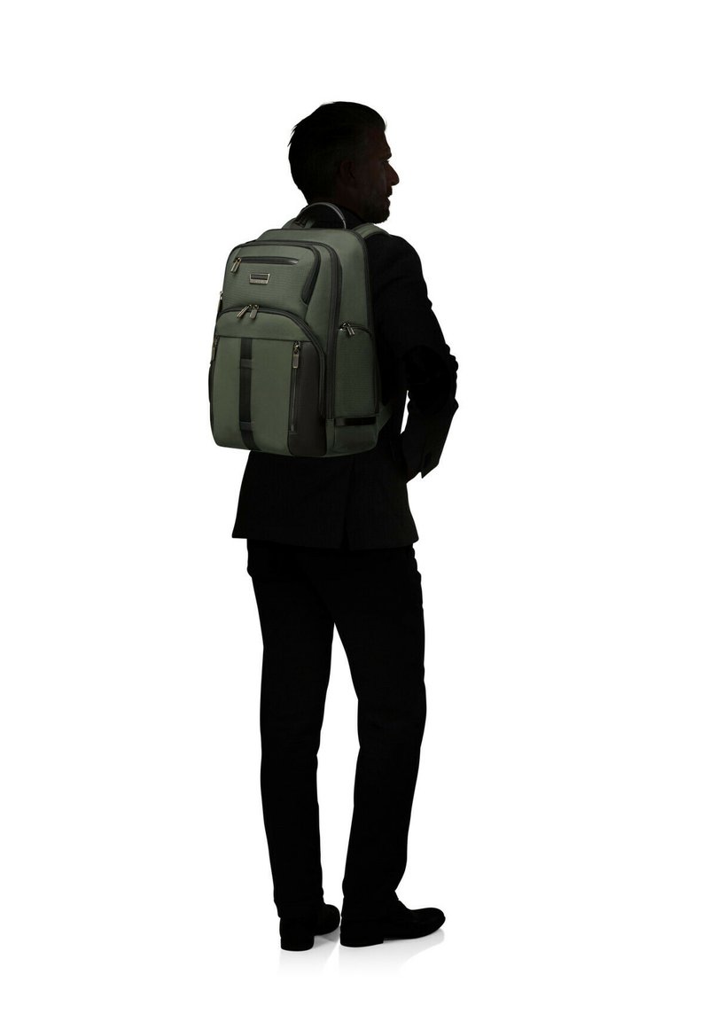 Dunkelgrüner Rucksack mit mehreren Außentaschen, strukturiertem Stoff und schwarzen Akzenten. Wird auf dem Rücken einer Person in einem Anzug getragen.