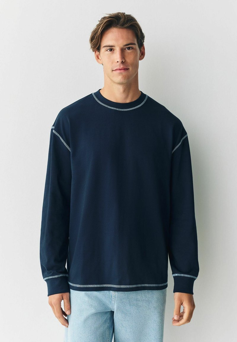 Sweatshirt lung cu mânecă lungă de culoare bleumarin, cu cusături deschise la culoare, gât rotund și o croială lejeră. Purtați-l cu blugi de culoare albastră deschisă.