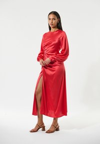 Rotes Satin-Kleid mit langen, gepufften Ärmeln, Raffung in der Taille und einem Oberschenkel-spalt. Getragen mit braunen Riemchensandalen.