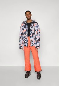 Muž oblečený v černém tričku, mnohobarevné bundě se abstraktním vzorem na lyže, zářivě oranžových lyžařských kalhotách a černých lyžařských botách, stojící na jednobarevném pozadí.