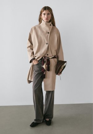 Frau trägt beigefarbenen hochgeschlossenen Mantel mit Knöpfen, dunkle weit geschnittene Jeans, schwarze Schuhe, hält braune Tasche, mit gemustertem Schal um die Taille gebunden.