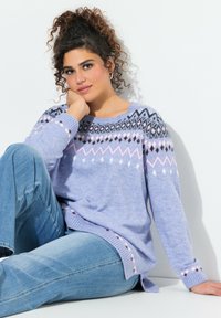 Maglione lilla chiaro con motivi geometrici in nero, bianco e rosa. Tessuto morbido, polsini a coste e spacchi laterali, abbinato a jeans blu.