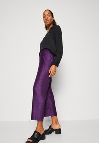 Černá blusa s dlouhým rukávem a límečkem, spárovaná s fialovými, texturovanými kalhotami s širokými nohavicemi a černými, otevřenými block-heel mule střevíci.