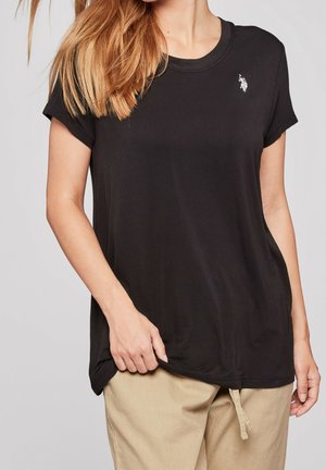 Donna che indossa una T-shirt nera a maniche corte con un piccolo logo bianco sul petto e pantaloni beige con coulisse, tenendo l'orlo della maglietta.