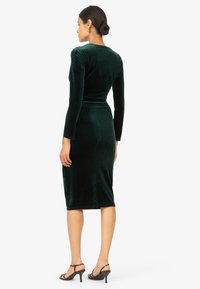Groene fluwelen jurk met lange mouwen, aansluitend ontwerp, knielang, gladde textuur, met een subtiele glans, gecombineerd met zwarte strapless hakken.