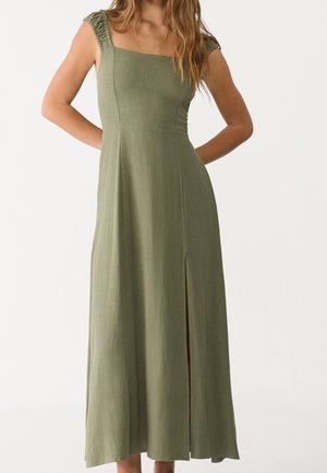 Woman wearing sleeveless olive green maxi dress with square neckline and front slit, posing with hands behind back.