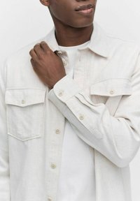 Lys beige knappet skjorte lavet af tekstureret stof, med to brystlommer og op-rullede manchetter. Båret over en hvid t-shirt.