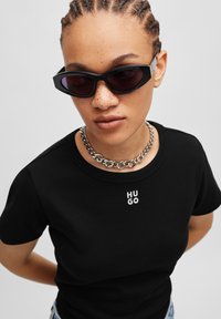 T-shirt en coton noir avec un logo "HUGO" blanc sur le devant, associé à des lunettes de soleil noires angulaires et un collier en chaîne argenté.