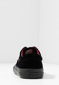 Mustat mokkasiinijalkineet, joissa on pyöreä kärki, kuviollinen punainen vuori ja kumipohja, jossa on hillitty logo painettuna kantapään kohdalle.