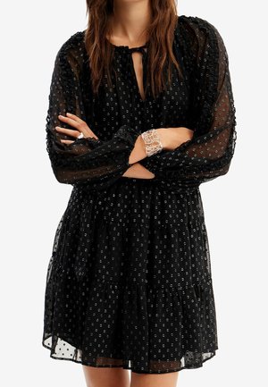 Black long-sleeve sheer dress with small polka dots, ruffled tiers, and keyhole neckline, worn with silver cuff bracelet and ring.