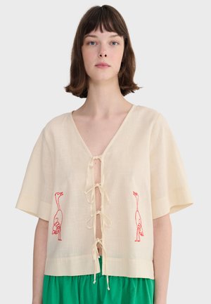 Femme portant un chemisier crème à manches courtes avec des liens devant et des oiseaux brodés rouges, associé à un pantalon vert vif.