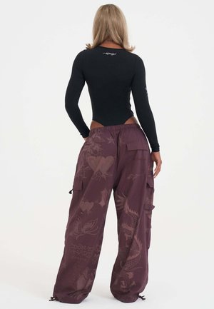 Vrouw met schouderlang blond haar draagt een zwarte top met lange mouwen en losse paarse cargobroek met tatoeage-geïnspireerde prints, kijkt weg van de camera.