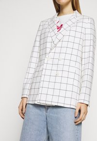 Blazer blanc à double boutonnage avec un motif à carreaux noirs, présentant des revers notched, deux boutons et des poches latérales, associé à un jean bleu clair.