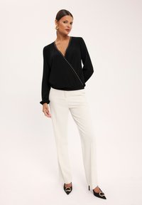 Czarna bluzka z głębokim dekoltem w kształcie V, ozdobiona metalicznymi akcentami po bokach, zestawiona z białymi eleganckimi spodniami i czarnymi szpilkami.