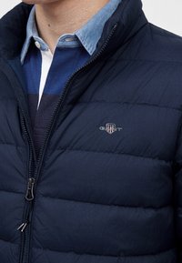 Homme portant une veste matelassée bleu marine avec une fermeture éclair, superposée sur une chemise bleue et blanche à col et un pull rayé bleu marine.