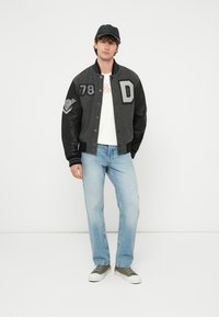 Mladý muž s čiernou čiapkou, sivou a čiernou univerzitnou bundou s nášivkami, bielym tričkom, svetlomodrými džínsami a sivými teniskami stojaci na bielom pozadí.