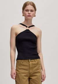Zwarte geribbelde haltertop met een driehoekige vorm, voorzien van een gouden ringdetail bij de halslijn en dunne bandjes. Draagt met beige broeken.