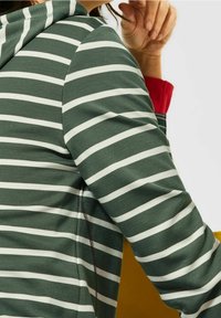 Persona che indossa un hoodie a righe verdi e bianche con un polsino rosso visibile, appoggiando la mano vicino al viso su uno sfondo neutro.