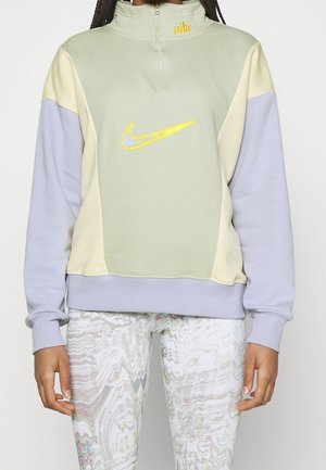 Person iført en Nike pastel color-block pullover med gul logo og mønstrede hvide leggings, der står mod en ensfarvet baggrund.