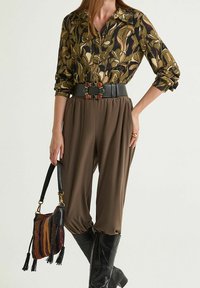 Bluse mit floralem Muster in Dunkelgrün und Braun, kombiniert mit locker sitzenden braunen Hosen, einem breiten schwarzen Gürtel mit farbenfrohen Akzenten und schwarzen Stiefeln.