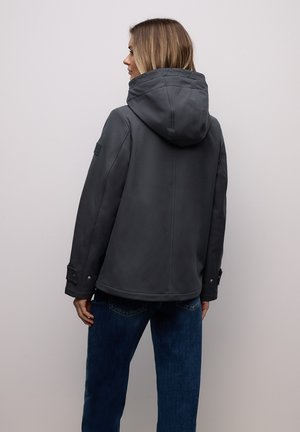 Femme portant une veste à capuche gris foncé et un jean bleu, debout de dos contre un mur clair uni.